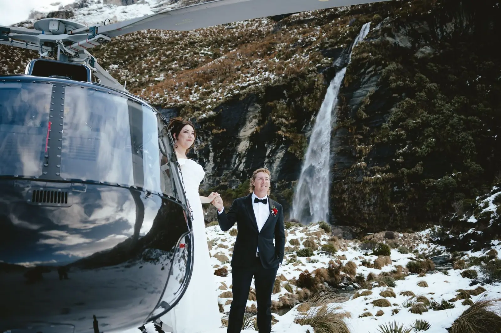 Helicopter landing beside a couple at Earnslaw Burn with waterfalls and snow behind