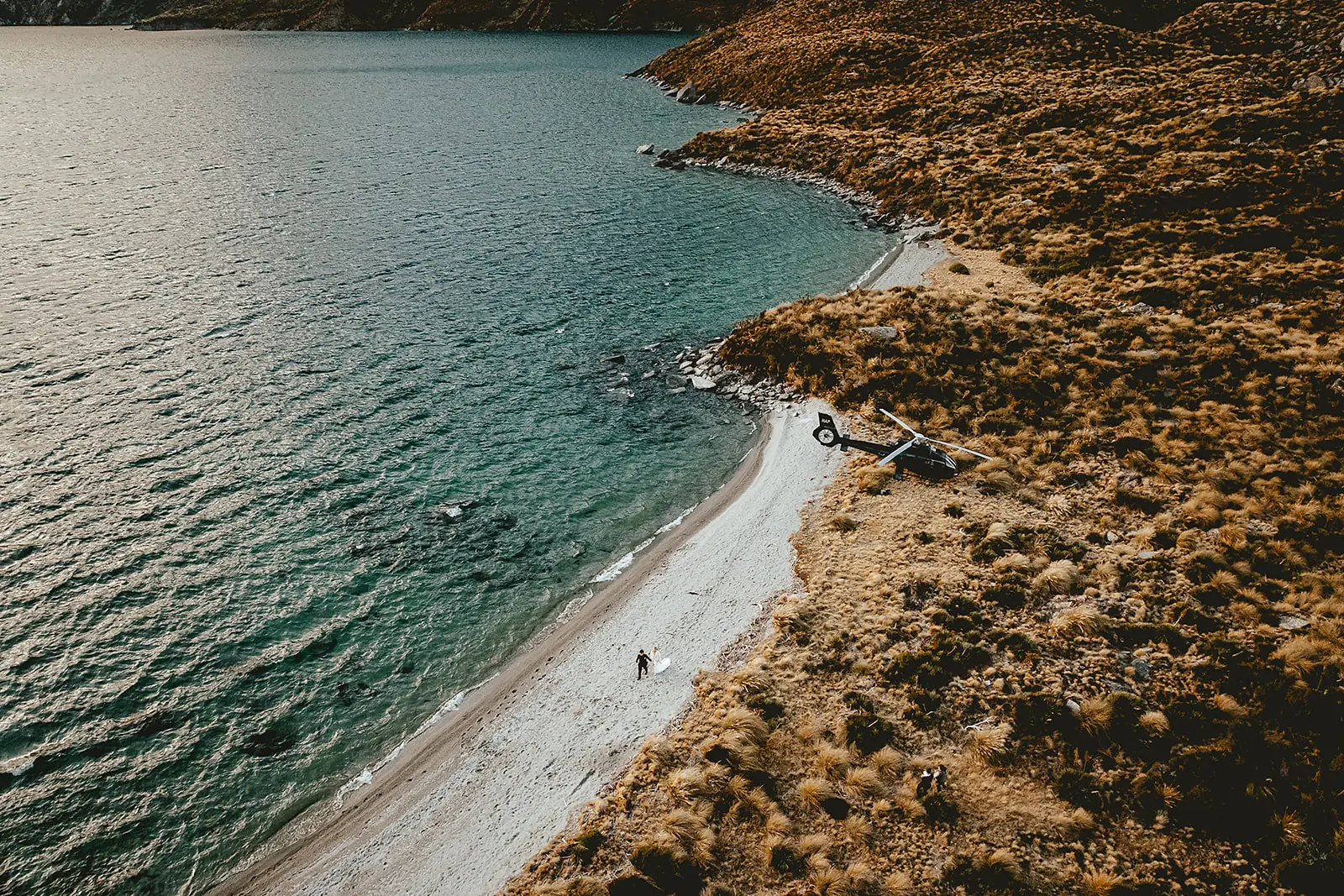 Aerial view of a couple and helicopter on the shore of a turquoise alpine lake near Queenstown
