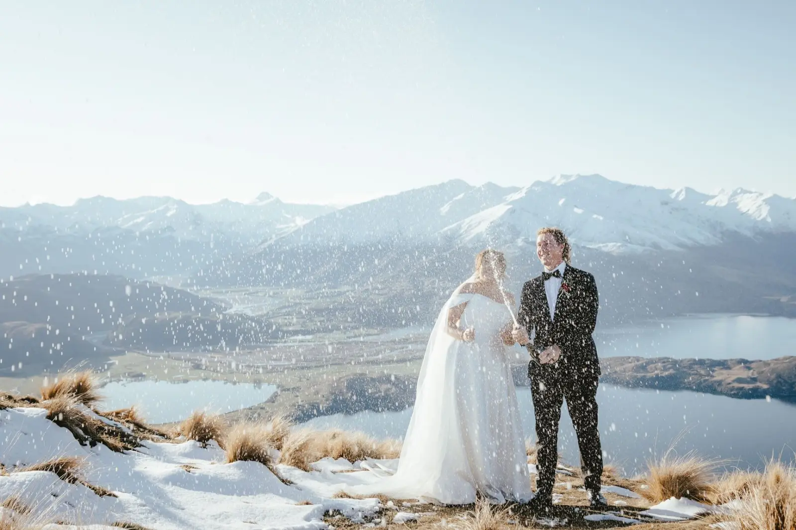 Couple spraying champagne on a snowy peak with mountains behind