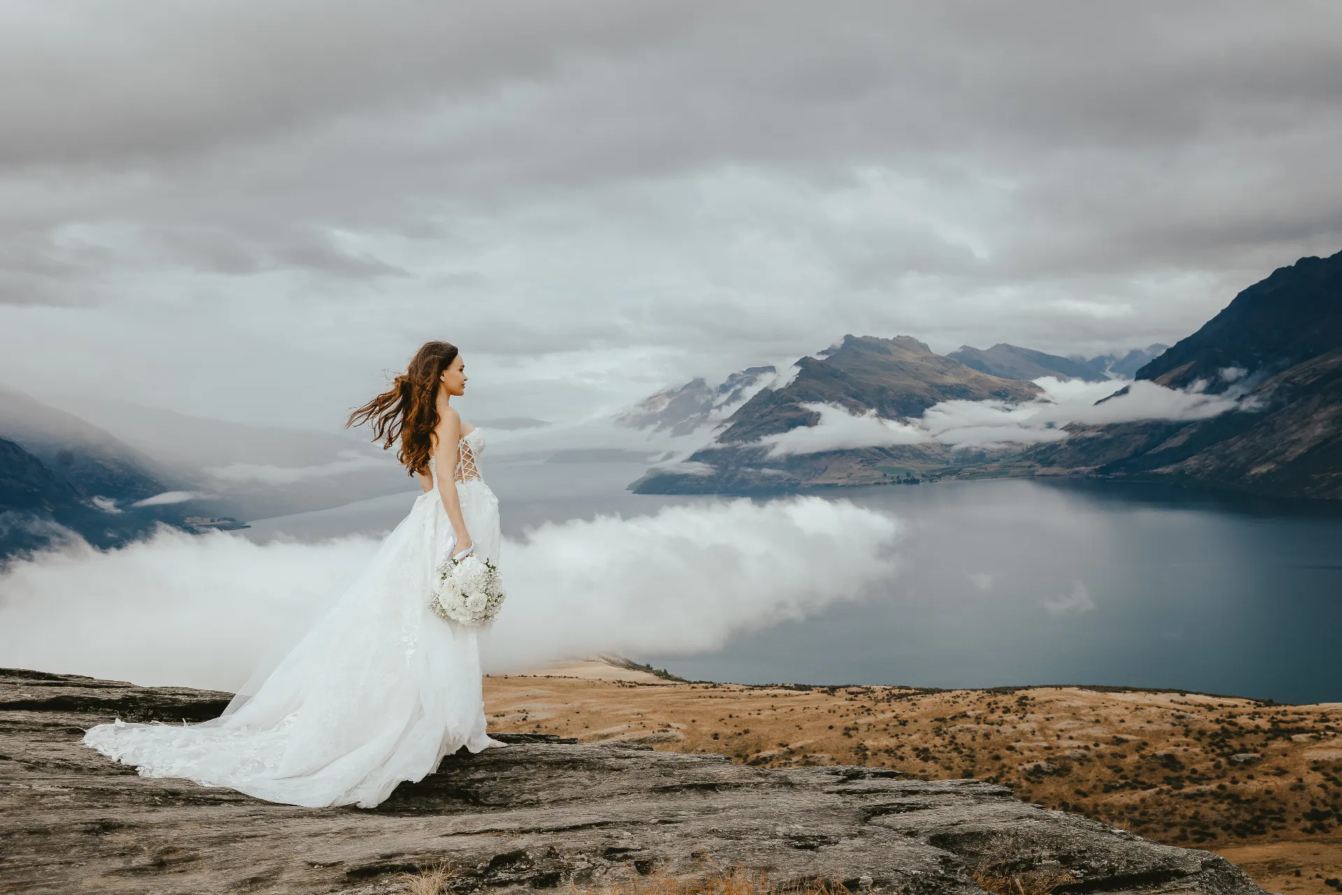 Bride at an elevated drive-up viewpoint above Queenstown with mountains behind her