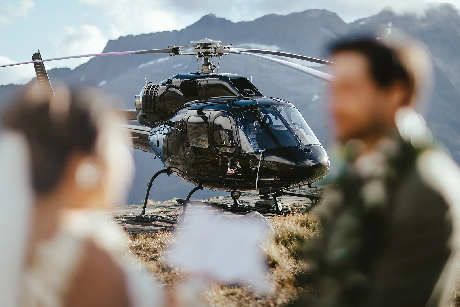 Bride and groom in the foreground as a helicopter lifts off beside an alpine lake in the Remarkables