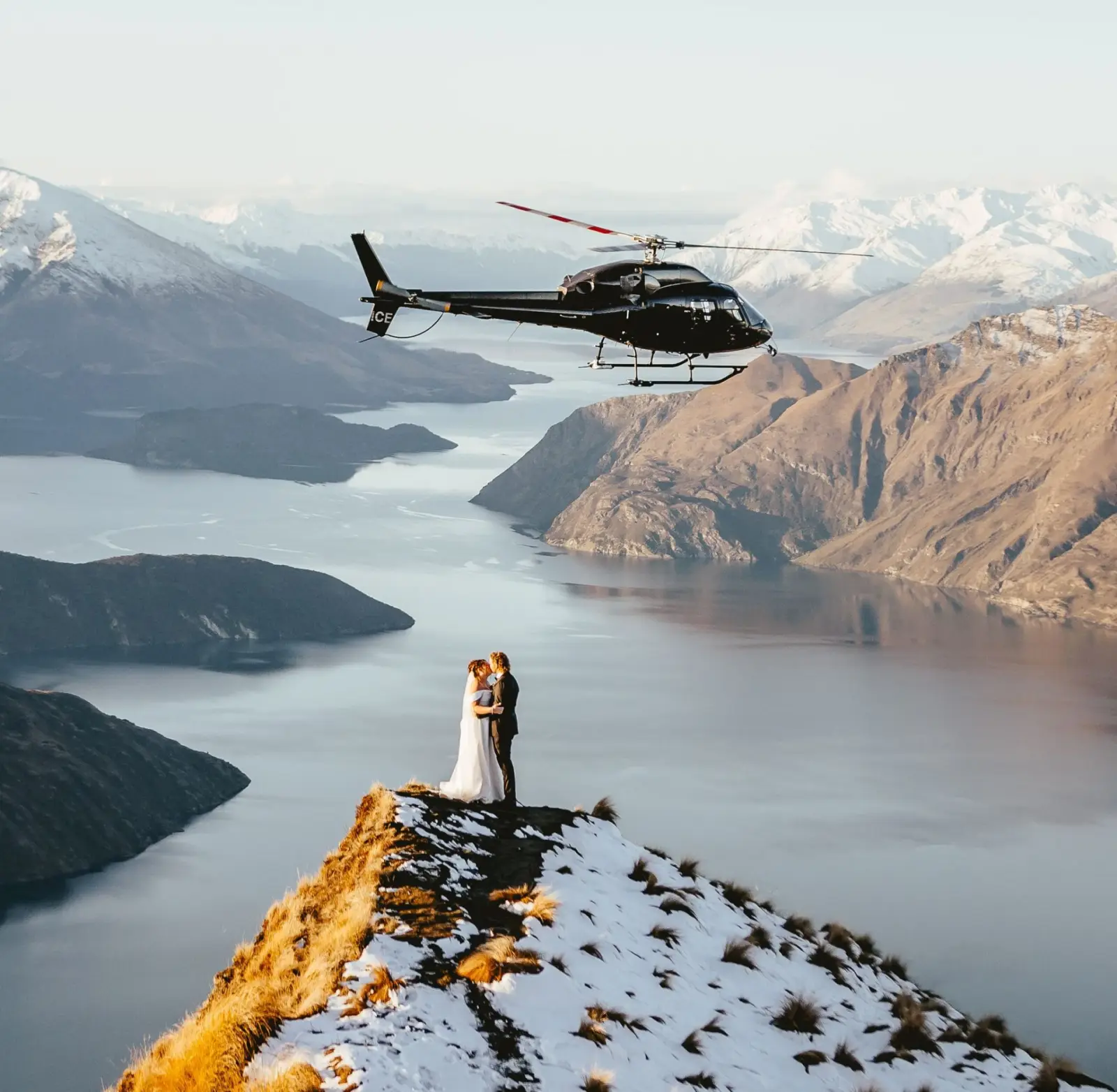 Couple kissing on the snow ridge at Coromandel Peak as the helicopter hovers above the lake